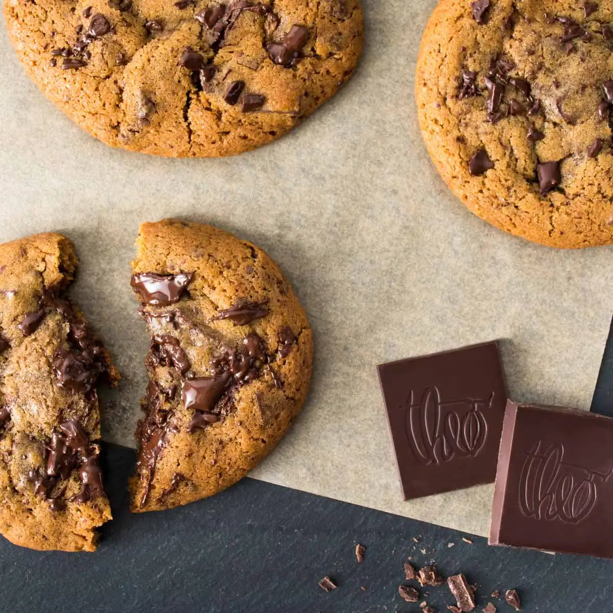 Chocolate chip cookies with chunks of chocolate on a textured surface, featuring a Theo brand logo.